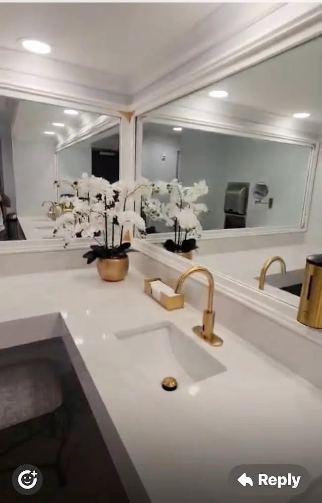 Modern bathroom vanity with white countertop, dual gold faucets, white orchid plants in gold planters, and large mirrors with recessed lighting