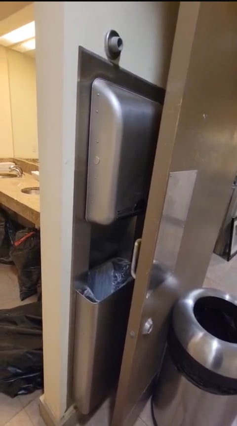 Tall stainless steel robot mounted on white wall pillar in indoor office setting with equipment visible