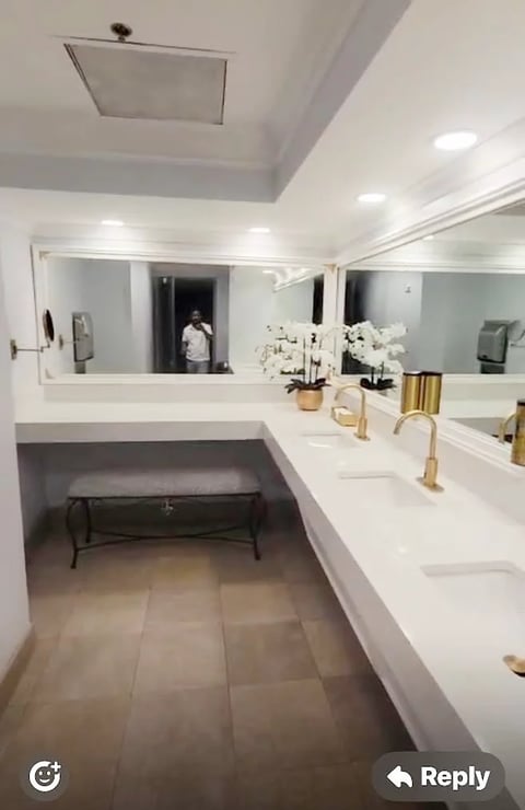 Modern luxury bathroom with double vanity, illuminated mirrors, white countertops, potted orchids, wooden accents, and dark tile flooring