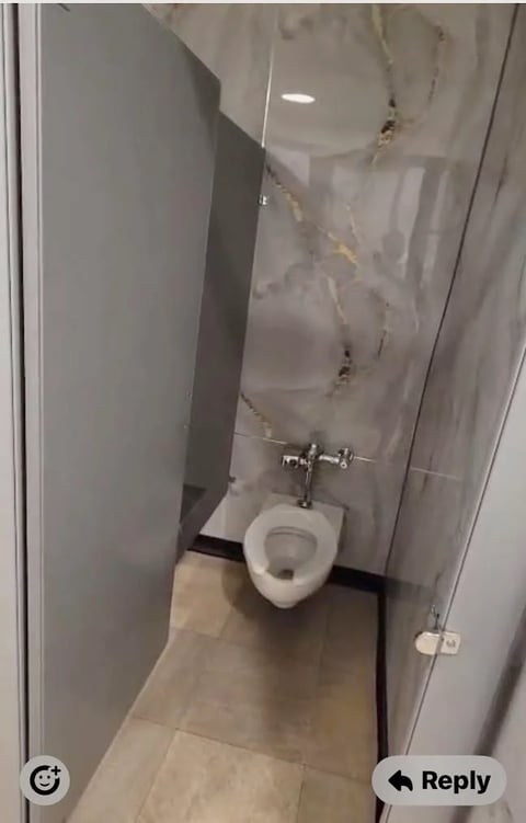 Small bathroom stall with white toilet, tile walls showing water damage stains, and wooden flooring